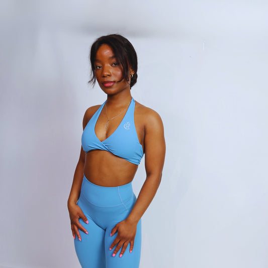 A female model wearing Spa blue halter sports bra with a twist front design, matching blue leggings. 